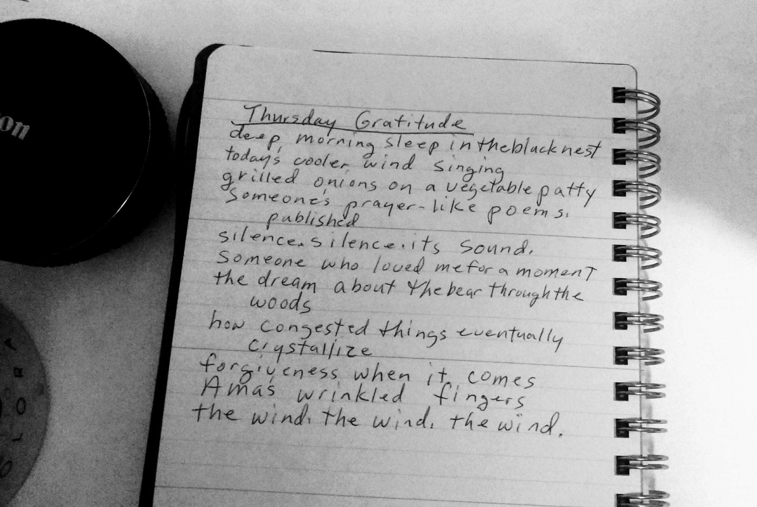 Black and white photo of notebook with a gratitude poem, a canon lengs, and coffee cup.
